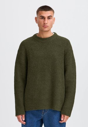 Hombre de pelo corto lleva un jersey verde oliva texturizado y vaqueros azules, de pie frente a un fondo liso y claro, mirando hacia adelante.