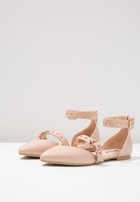 Zapatos planos de color rosa pálido con puntera afilada, correa alrededor del tobillo con tachuelas doradas y cierre de hebilla; textura de cuero suave.