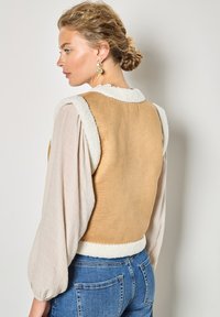 Weste in Tan ohne Ärmel mit weißem Lammfellbesatz und Kragen, getragen über einer hellen strukturierten Bluse. Hat ein tailliertes Design und Pastellfarben.