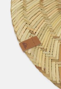 Pyöreä kudottu olkamatto luonnollisessa värimaailmassa, jossa on ruskea nahkaleima ommeltu yhdelle reunalle, esittelee logon.