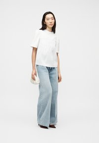 Γυναίκα που φοράει λευκό μπλουζάκι με κοντά μανίκια και φαρδιά ανοιχτό μπλε τζιν, κρατώντας μικρή λευκή τσάντα χειρός, στέκεται μπροστά σε λευκό φόντο.