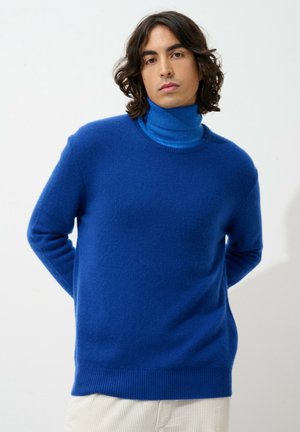 Royalblauer Rollkragenpullover aus weichem Strick. Mit lockerem Schnitt, gerippten Säumen und Bündchen. Das Model hat die Hände hinter dem Rücken und trägt cremefarbene Hosen.