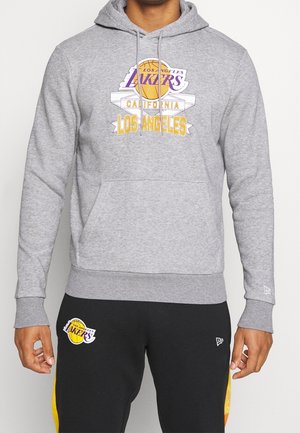 Osoba ubrana w szarą bluzę z kapturem Los Angeles Lakers z logo koszykówki oraz czarne spodnie z emblematem Lakers i żółtymi paskami po bokach.