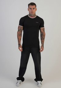 Black fitted t-shirt with short sleeves and white trim, paired with dark jeans. Model stands neutrally, showcasing tattooed arms and sneakers.