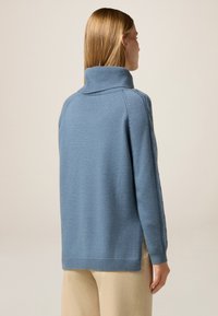 Niebieski sweter z dzianiny o wysokim kołnierzu, długich rękawach, ściągaczach przy mankietach i u dołu, z bocznymi szczelinami i gładką teksturą. Stylizowany na beżowych spodniach.