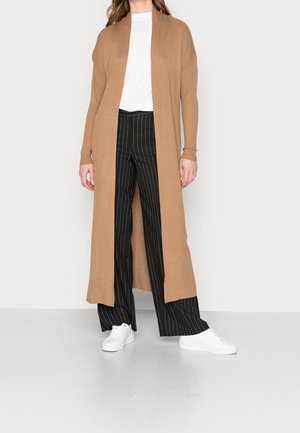 Femme portant un long cardigan couleur camel, un haut blanc, un pantalon large noir à fines rayures, et des baskets blanches, debout devant un fond clair uni.