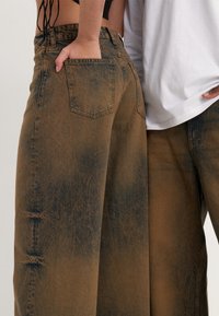 Hnědé džíny s širokými nohavicemi a vysokým pasem, s vybledlou texturou a tmavými akcenty. Mají zadní kapsy a volný střih. Částečně vidět dva modely.