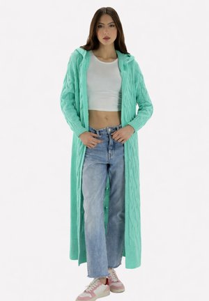 Joven mujer con cárdigan largo de punto azul claro, top corto blanco, vaqueros azul claro y zapatillas rosas y blancas de pie contra un fondo blanco.