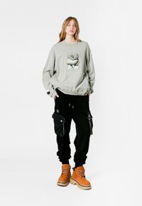 Sudadera gris con un gráfico de dibujo animado, combinada con pantalones cargo negros con bolsillos laterales y botas de cordones naranjas con suela gruesa.