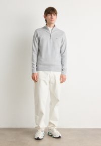 Pulover gri cu fermoar la gât, cu tricot texturat, asortat cu pantaloni albi cu croială lejeră și adidași multicolori. Stând în fața unui fundal neutru.