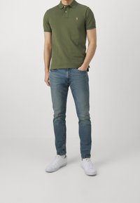 Polo shirt verde oliva con due bottoni, abbinato a jeans in denim azzurro chiaro e sneakers bianche. Il polo presenta un piccolo logo ricamato.