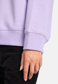 Sudadera lila con puños acanalados; mano descansando sobre pantalones de pana negra. Textura suave, diseño simple y corte relajado.