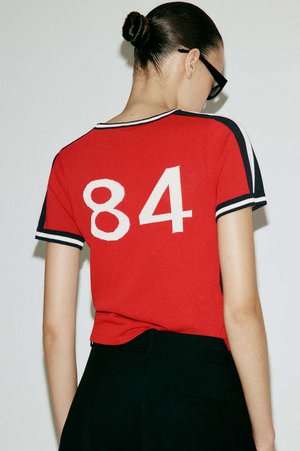 Mujer con el cabello recogido en un moño, lleva gafas negras y una camiseta roja de manga corta con el número 84 blanco en la espalda, combinada con pantalones negros.