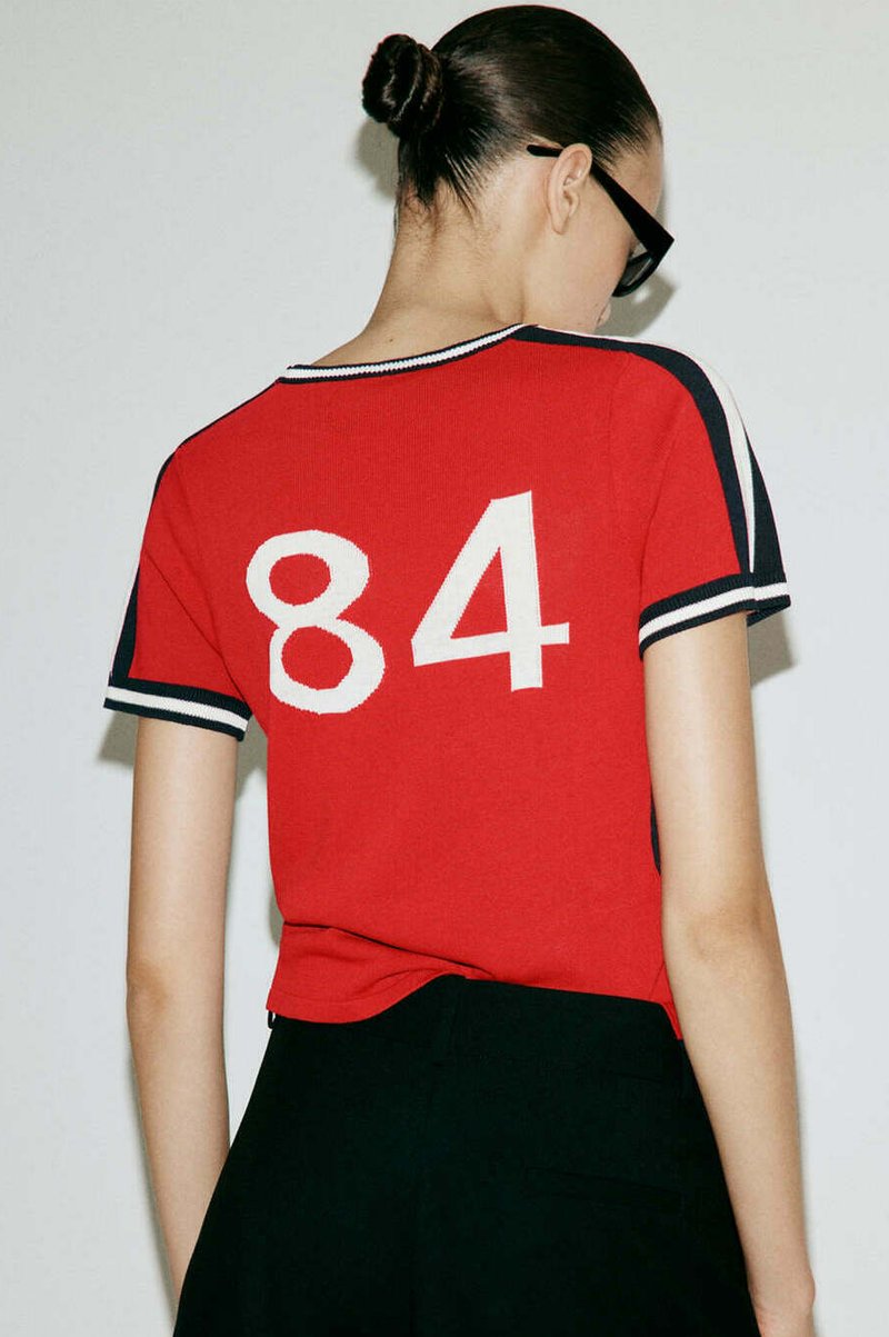 Femme avec les cheveux en chignon portant des lunettes noires et un t-shirt rouge à manches courtes avec le numéro blanc 84 dans le dos, associé à un pantalon noir.