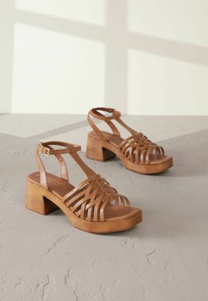 Rjave usnjene sandale na blok peti z tkanimi trakovi, zapenjanjem okrog gležnja in gladko leseno platformo. Teksturirana površina z mat zaključkom.