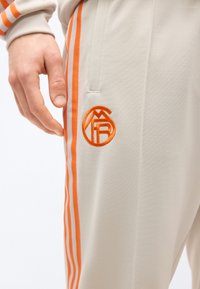 Ruka spočívajúca pri béžových nohaviciach s oranžovými zvislými pruhmi a vyšívaným kruhovým logom blízko vrecka.