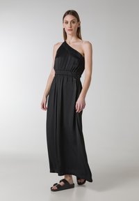 Sort, one-shoulder maxi kjole med elastisk talje. Glat, letvægtsstof. Modellen har sorte flade sandaler på.