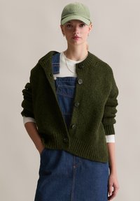 Gilet tricoté vert foncé avec de grands boutons, porté par-dessus une chemise blanche à manches longues et des salopettes en denim bleu. Texture douce et coupe ample.