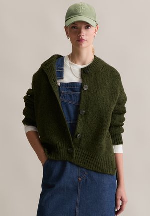 Donkergroene gebreide cardigan met grote knopen, gedragen over een witte lange mouwen shirt en blauwe denim tuinbroek. Zachte textuur en losse pasvorm.