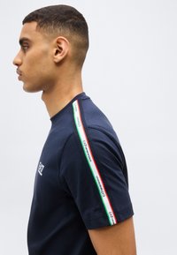 Νέος άνδρας με μπλε ναυτικό μπλουζάκι με ταινία με κόκκινες, λευκές και πράσινες ρίγες στο μανίκι με το λογότυπο EA7 Emporio Armani, κοιτάζοντας προς τα αριστερά σε απλό φόντο.