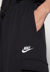 Svarta joggers gjorda av mjukt material, med en sidoficka och en framträdande vit Nike-logga broderad på låret.