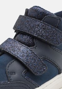 Sneaker blu scuro con due fascette in Velcro glitterate, materiale simile alla pelle liscio e suola testurizzata con accenti bianchi.