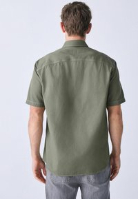 Camisa de manga corta en tejido verde oliva, con un cuello estructurado, superficie texturizada, dobladillo curvado y un diseño de corte relajado.