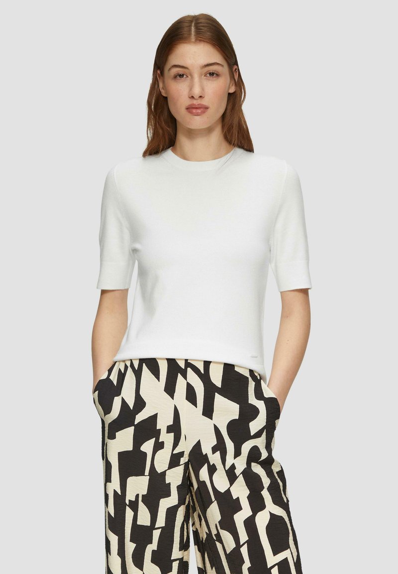 Haut en tricot blanc à manches courtes avec un col rond, associé à un pantalon large à motifs noirs et crème. Détails de tissu texturé visibles.