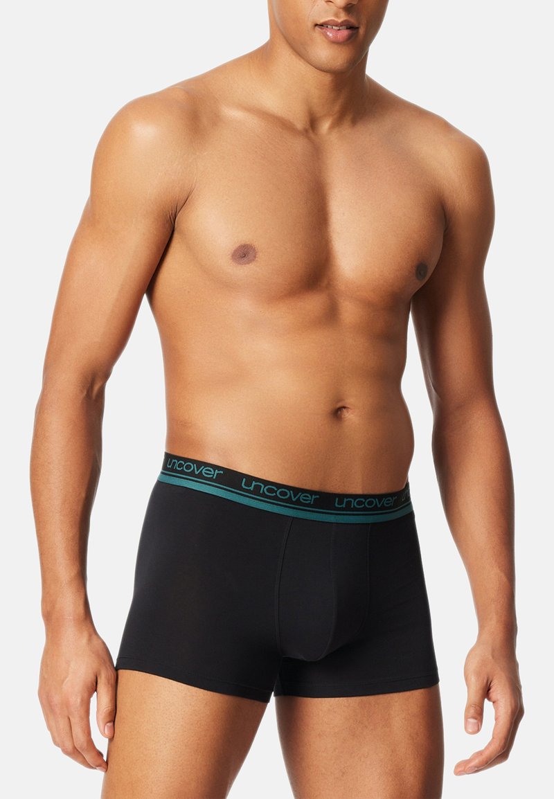 Black boxer briefs made of soft fabric, featuring a turquoise elastic waistband with “uncover” brand text. Fitted design with no patterns.