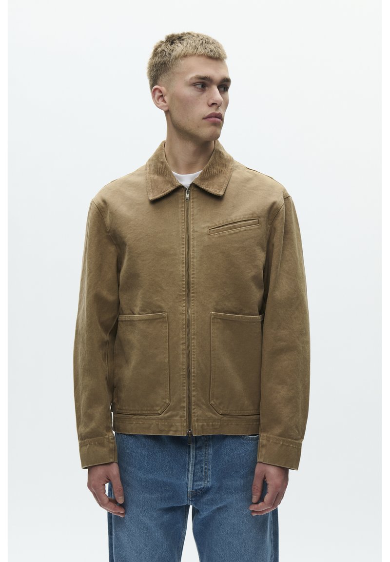 Ruskea vetoketjullinen takki, jossa kaulus, kaksi etutaskua ja rintatasku. Valmistettu teksturoidusta kankaasta. Yhdistetty sinisiin farkkuihin.