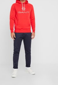 Sweat à capuche rouge avec logo sur la poitrine, doté d'une poche avant et de cordons blancs. Associé à un pantalon slim bleu marine et des baskets blanches.