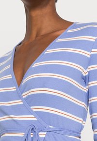 Blå omslagstopp med vita och orange horisontella ränder. Har en v-ringning och en knytning i midjan. Ribbad textur.