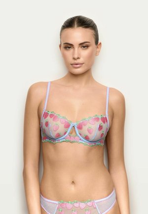 Donna con capelli raccolti che indossa un reggiseno e slip lilla trasparenti ricamati con fragole rosa e bordi verdi, su sfondo semplice.