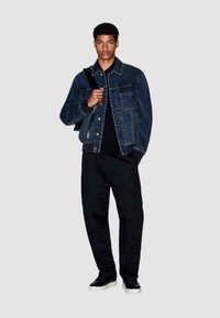 Donkere blauwe spijkerjas met een knoopsluiting aan de voorkant, kraag en twee borstzakken, gecombineerd met zwarte broek en sneakers met witte zolen.