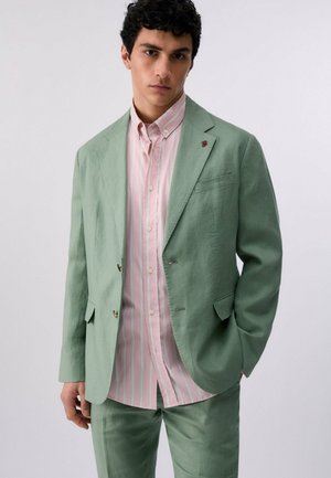 Hombre con un traje verde claro y una camisa abotonada con rayas rosas y blancas, de pie frente a un fondo claro y sencillo.