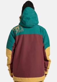 Chaqueta colorida con una sección superior verde azulado, una parte central burdeos y acentos amarillos, con un logo prominente en la parte trasera. Hecha de un material duradero.