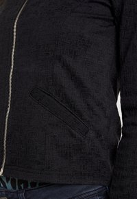 Černá bunda na zip s texturovaným vzorem, boční kapsou a přiléhavým střihem. Vyrobena z měkké, pružné tkaniny s hladkým zipem.