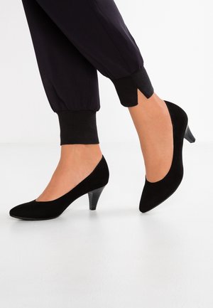 Czarne zamszowe szpilki z wąskim noskiem i niskim, skośnym obcasem. Noszone z czarnymi spodniami dresowymi z prążkowanymi mankietami.