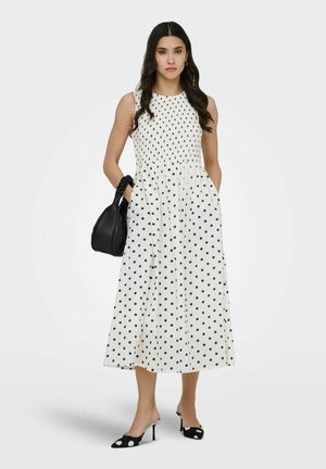 Kvinde iført hvid ærmeløs kjole med sorte polkaprikker, sorte polkaprikker-hæle og holder en sort håndtaske med rynket håndtag.