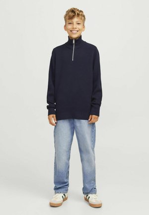 Pull à col zippé marine en tissu tricoté, coupe décontractée, associé à un jean bleu clair et des baskets blanches avec des accents verts.