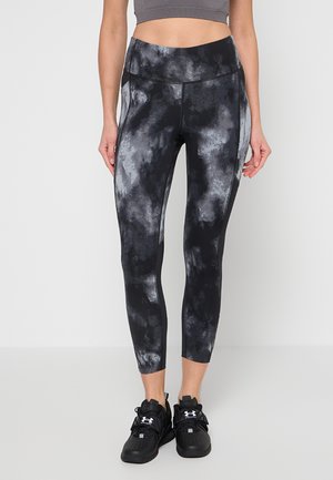 Kobieta w czarno-szarych legginsach z wysokim stanem i wzorem tie-dye oraz w czarnych sportowych butach Under Armour, stojąca na jednolitym tle.