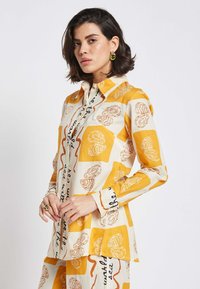 Long-sleeve shirt in cream and yellow featuring an abstract rose pattern and handwritten text, with a collared neck and button-down front.