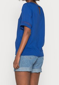 Top blu corto a maniche corte con schiena a chiave, caratterizzato da un pizzo sulle maniche. Abbinato a pantaloni corti in denim arrotolati.