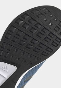 Semelle extérieure noire texturée en caoutchouc d'une chaussure avec des crampons en forme de pyramide et une partie de la tige en mesh bleu visible.