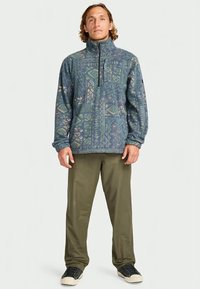 Man staat in een blauwe fleece met een patroon en olijfgroene broek met zwarte sneakers tegen een effen achtergrond.