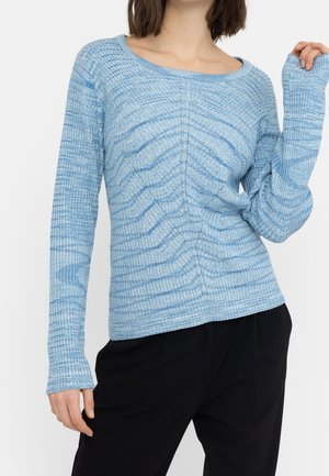 Femme portant un pull en maille à manches longues, bleu clair à motifs, et un pantalon noir, posant avec une main levée près de la tête.