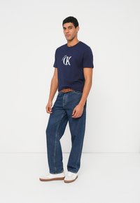 Tumši zila kokvilnas T-krekls ar baltu logotipu, kombinēts ar brīvā piegriezuma zilām džinsa biksēm un baltām sporta kurpēm ar brūnām zolēm un akcentiem.