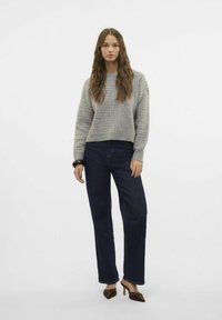Suéter de punto texturizado gris claro con un diseño cropped, combinado con vaqueros de pierna ancha oscuros. La modelo está de pie con una pulsera en una muñeca.