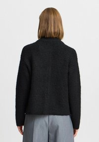 Čierny pletený sveter s textúrovaným vzorom, s mierne oversized strihom a s vysokým golierom, nosený cez šedé nohavice.