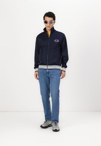 Giacca nera con zip e polsini a strisce bianche, logo sul petto, abbinata a jeans azzurri e sneakers grigie. Uomo che indossa occhiali da sole.